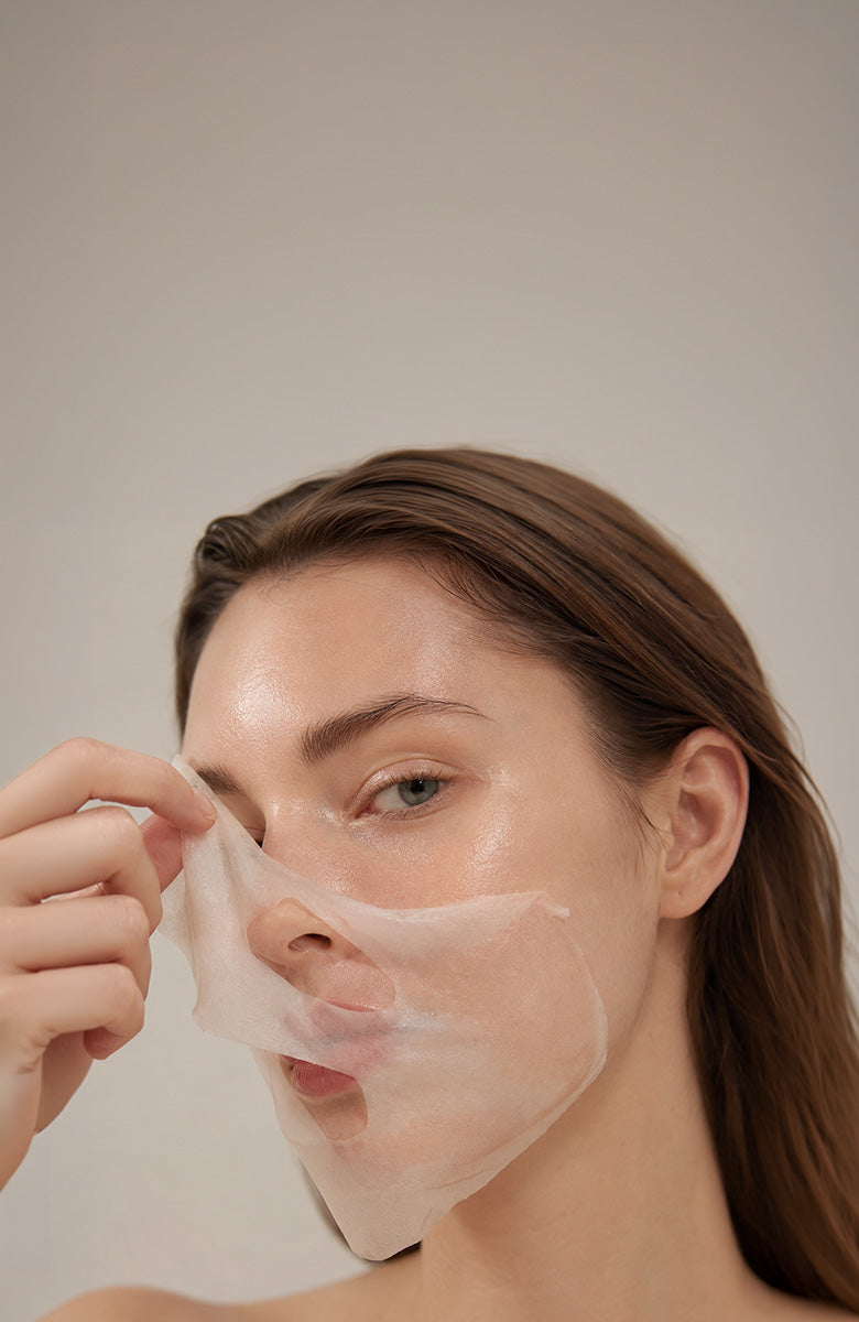 A woman with brown hair is shown from the shoulders up, peeling a translucent white sheet mask off her face against a plain light beige background.